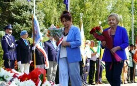 Dwie kobiety składające wiązankę biało-czerwonych kwiat&oacute;w pod pomnikiem. W tle poczty sztandarowe.