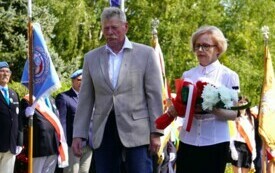 Dwoje ludzi składających wiązankę biało-czerwonych kwiat&oacute;w pod pomnikiem. W tle poczty sztandarowe.