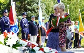 Dwie kobiety składające wiązankę biało-czerwonych kwiat&oacute;w pod pomnikiem. W tle poczty sztandarowe.