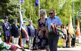 Dwoje ludzi składających wiązankę biało-czerwonych kwiat&oacute;w pod pomnikiem. W tle poczty sztandarowe.