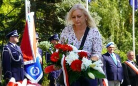 Kobieta składająca wiązankę biało-czerwonych kwiat&oacute;w pod pomnikiem. W tle poczty sztandarowe.