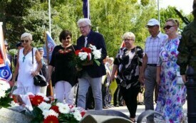 Grupa ludzi składających wiązanki biało-czerwonych kwiat&oacute;w pod pomnikiem.
