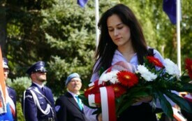 Kobieta trzymająca wiązanę biało-czerwonych kwiat&oacute;w pod pomnikiem. W tle poczty sztandarowe.