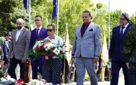 Grupa ludzi składających wiązanki biało-czerwonych kwiat&oacute;w pod pomnikiem. W tle poczty sztandarowe.