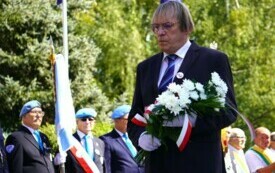 Mężczyzna składający wiązankę biało-czerwonych kwiat&oacute;w pod pomnikiem. W tle poczty sztandarowe.