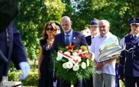 Grupa ludzi składających wiązanki biało-czerwonych kwiat&oacute;w pod pomnikiem.