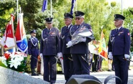 Grupa mundurowych składających wiązanki biało-czerwonych kwiat&oacute;w pod pomnikiem.