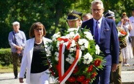 Elegancko ubrani kobieta i mężczyzna, przed nimi strażnik miejski w wieńcem biało-czerwonych kwiat&oacute;w.