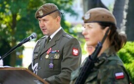 Żołnierz przemawiający do mikrofonu przy m&oacute;wnicy, na pierwszym planie kobieta w mundurze wojskowym. W tle zieleń drzew i krzew&oacute;w.