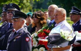 Grupa ludzi, część w mundurach, część elegancko ubranych. Niekt&oacute;rzy trzymają wiązanki biało-czerwonych kwiat&oacute;w