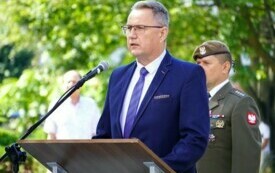 Mężczyzna przemawiający do mikrofonu przy m&oacute;wnicy, za nim stoi żołnierz. Wok&oacute;ł zieleń drzewi i krzew&oacute;w.