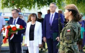 Grupa elegancko ubranych ludzi, kobieta i dw&oacute;ch mężczyzn. Jeden z mężczyzn trzyma wieniec biało-czerwonych kwiat&oacute;w. Na pierwszym planie kobieta w wojskowym mundurze, stojąca na warcie honowowej. 