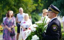 Grupa ludzi stojących w plenerze, na pierwszym planie strażnik miejski z wieńcem biało-czerwonych kwiat&oacute;w.