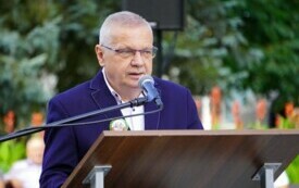 Mężczyzna przemawiający do mikrofonu przy m&oacute;wnicy, wok&oacute;ł zieleń drzew i krzew&oacute;w.