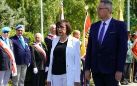 Dwoje elegancko ubranych ludzi, kobieta i mężczyzna stojący bok siebie. W tle poczty sztandarowe, ludzie z wieńcami kwiat&oacute;w.