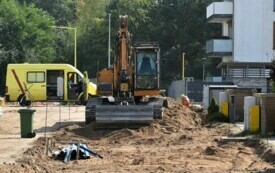 Plac budowy drogi, na pierwszym planie pracująca koparka. Po prawej stronie zabudowa wielorodzinna