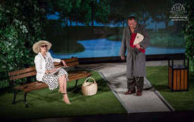 Zdjęcie ze spektaklu. Na scenie aranżacja parkowej alejki, na trawniku stoi ławeczka, za ławką "żywopłot". W tle wyświetlony widok z parkowej alejki. Na scenie mężczyzna i kobieta. Mężczyzna w piżamie i szlafroku stoi na środku alejki i trzyma w rękach ga