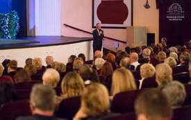 Mężczyzna w garniturze w sali widowiskowej przemawia do publiczności. W tle widać scenę, na kt&oacute;rej będzie odbywał się spektakl teatralny. Na pierwszym planie publiczność zgromadzona w sali, słuchająca przemawiającego mężczyzny.