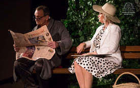 Zdjęcie ze spektaklu. Kobieta i mężczyzna siedzący na ławce, za nimi zieleń krzew&oacute;w. Na ziemi obok kobiety stoi kosz wiklinowy. Mężczyzna ubrany w piżamę i szlafrok, czyta gazetę. Kobieta siedzi obok, elegancko ubrana, w kapeluszu na głowie. Na kolanach t