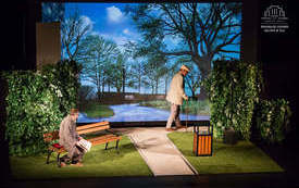 Zdjęcie ze spektaklu. Na scenie aranżacja parkowej alejki, obok kt&oacute;rej stoi kosz na śmieci i ławeczka, za ławką "żywopłot". W tle wyświetlony widok z parkowej alejki. Na scenie dw&oacute;ch mężczyzn: jeden siedzi na ławce w piżamie i szlafroku, trzyma w rękach g