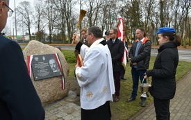 Grupa ludzi skupiona wok&oacute;ł pomnika - kamienia z tablicą pamiątkową. W centralnym miejscu ksiądz dokonuje poświęcenia pomnika. 