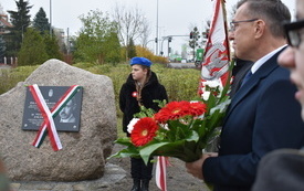 Kamień z tablicą pamiątkową, po kt&oacute;rej dw&oacute;ch stronach zwisają flagi Polska i Węgierska. Obok pomnika stoi harcerka w niebieskim berecie, obok niej poczet sztandarowy. Na pierwszym planie mężczyzna z wiązanką biało-czerwonych kwiat&oacute;w.