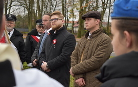 Grupa ludzi, wszyscy są odświętnie ubrani i skupieni, dw&oacute;ch mężczyzn w tle stoi w poczcie sztandarowym, dalej widać drzewa i zabudowania. Zdjęcie jest zrobione zza ramion innych ludzi stojących w grupie.