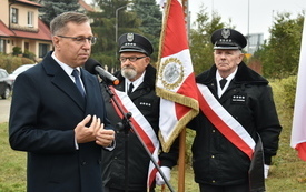 Na pierwszym planie mężczyzna przemawiający do mikrofonu. Za nim poczet sztandarowy, drzewa i zabudowania.