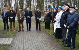 Grupa ludzi stojących na skwerze, wszyscy są odświętnie ubrani. W tle drzewa i ulica. Wśr&oacute;d zebranych są harcerze, ksiądz i wojskowy, jeden z mężczyzn trzyma w ręce bukiet biało-czerwonych kwiat&oacute;w. 