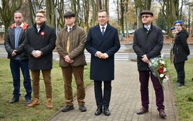 Pięciu mężczyzn stoi w rządzie na świeżym powietrzu, wszyscy są odświętnie ubrani. W tle drzewa na skwerze i jezdnia, po prawej stronie dwie harcerki w niebieskich beretach. Jeden z mężczyzn trzyma w ręce bukiet biało-czerwonych kwiat&oacute;w.  