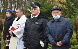 Cztery osoby stoją na świeżym powietrzu, wśr&oacute;d nich jest ksiądz, mundurowy oraz harcerka. Za nimi zielone i ż&oacute;łte liście na drzewach. 