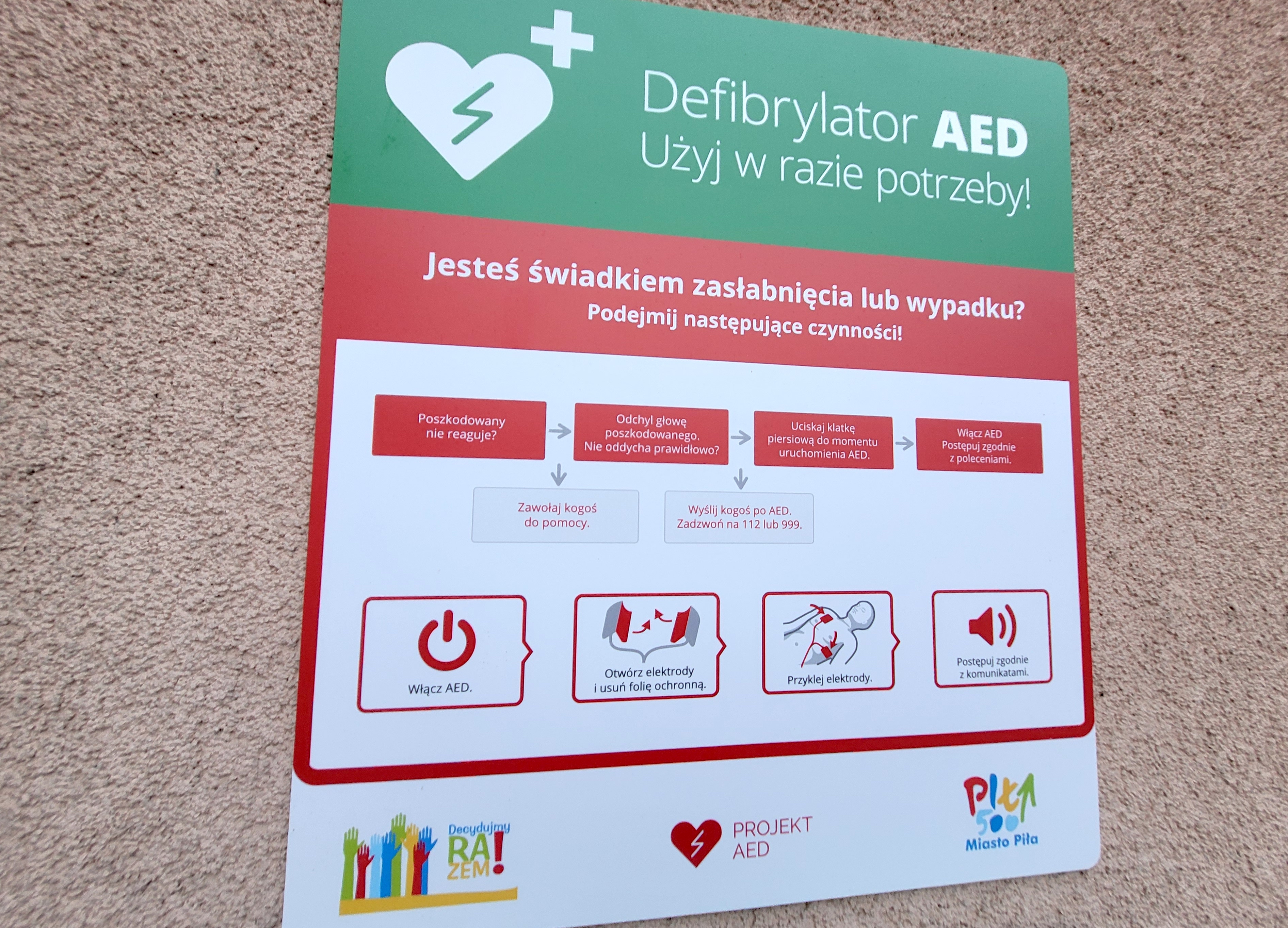 Tabliczka z informacją zawieszona obok defibrylatora AED. Treść tabliczki: Defibrylator AED Użyj w razie potrzeby. Jesteś świadkiem zasłabnięcia lub wypadku? Podejmij następujące czynności. Poszkodowany nie reaguje? Zawołaj pomoc, odchyl głowę poszkodowanego. Nie oddycha prawidłowo? Wyślij kogoś po AED. Zadzwoń na 112 lub 999. Uciskaj klatkę piersiową do momentu uruchomienia AED. Włącz AED i postępuj zgodnie z poleceniami. Pod instrukcją tekstową grafiki z dopiskiem: Włącz AED, Otwórz elektrody i usuń folię ochronną, przyklej elektrody, postępuj zgodnie z komunikatem. Na damym dole tabliczki logotyp Decydujmy razem, projekt AED, miasto piła. 