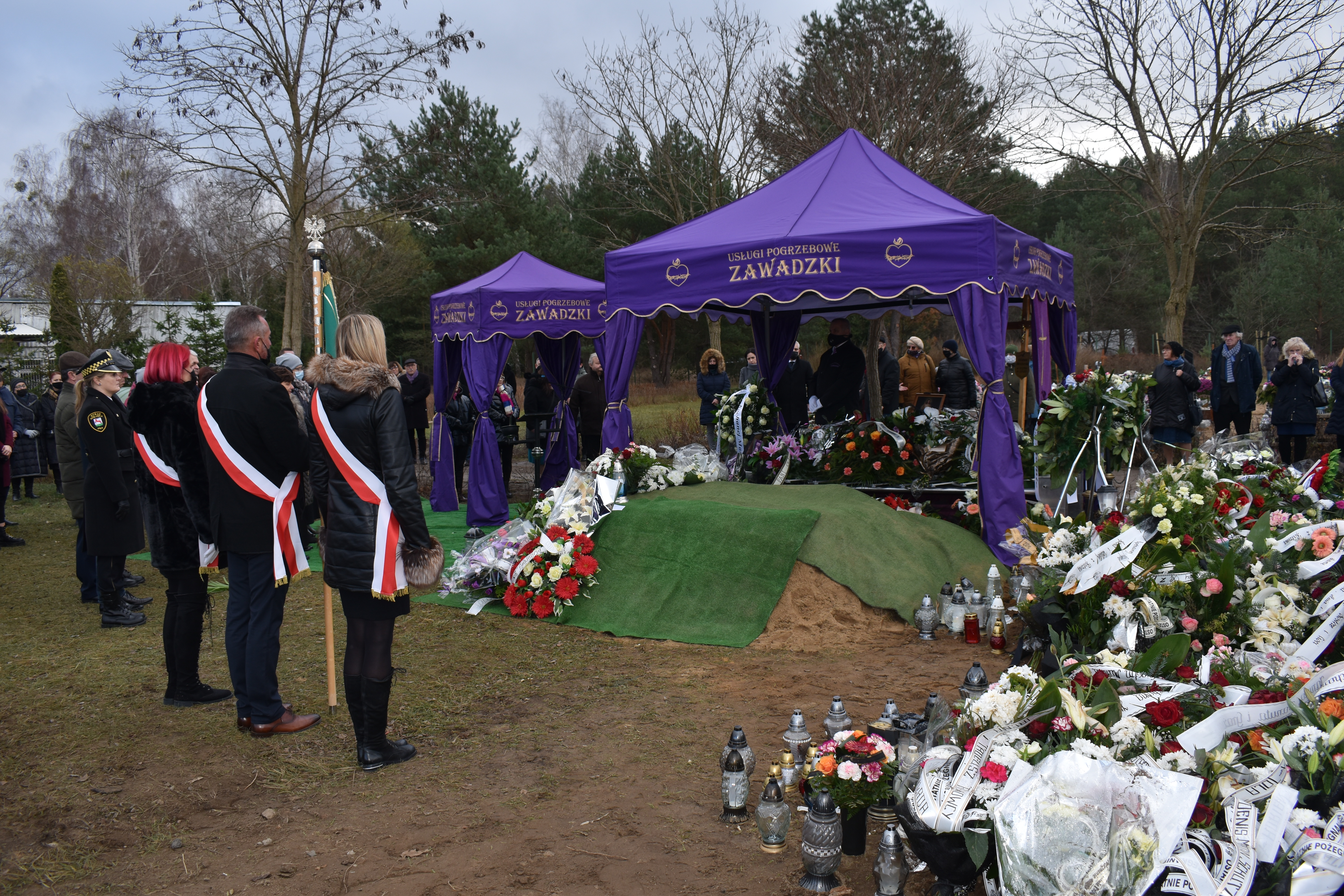 Zdjęcie ceremonii pogrzebowej na&nbsp;cmentarzu. Zgromadzeni ludzie stoją przy mogile. Na pierwszym planie poczet sztandarowy.