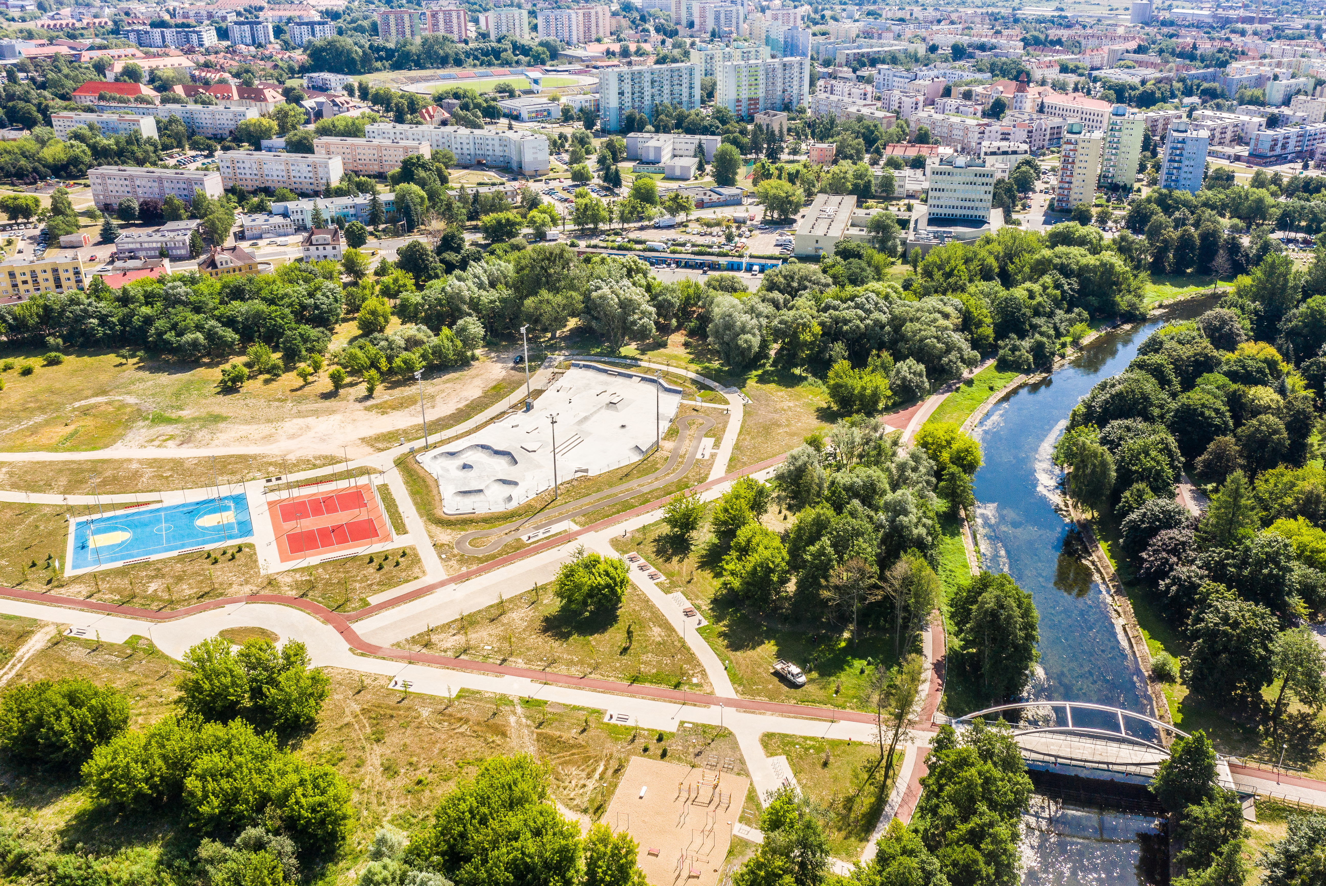 Zdjęcie fragmentu miasta z lotu ptaka. Na pierwszym planie kompleks obiektów sportowych i rekreacyjnych, otoczonych zielenią. Dwa boiska wielofunkcyjne, skateplaza, ścieżki rowerowe i piesze. Sportowe obiekty otacza zieleń. Po prawej stronie widać zakole rzeki. W tle budynki mieszkaniowe.