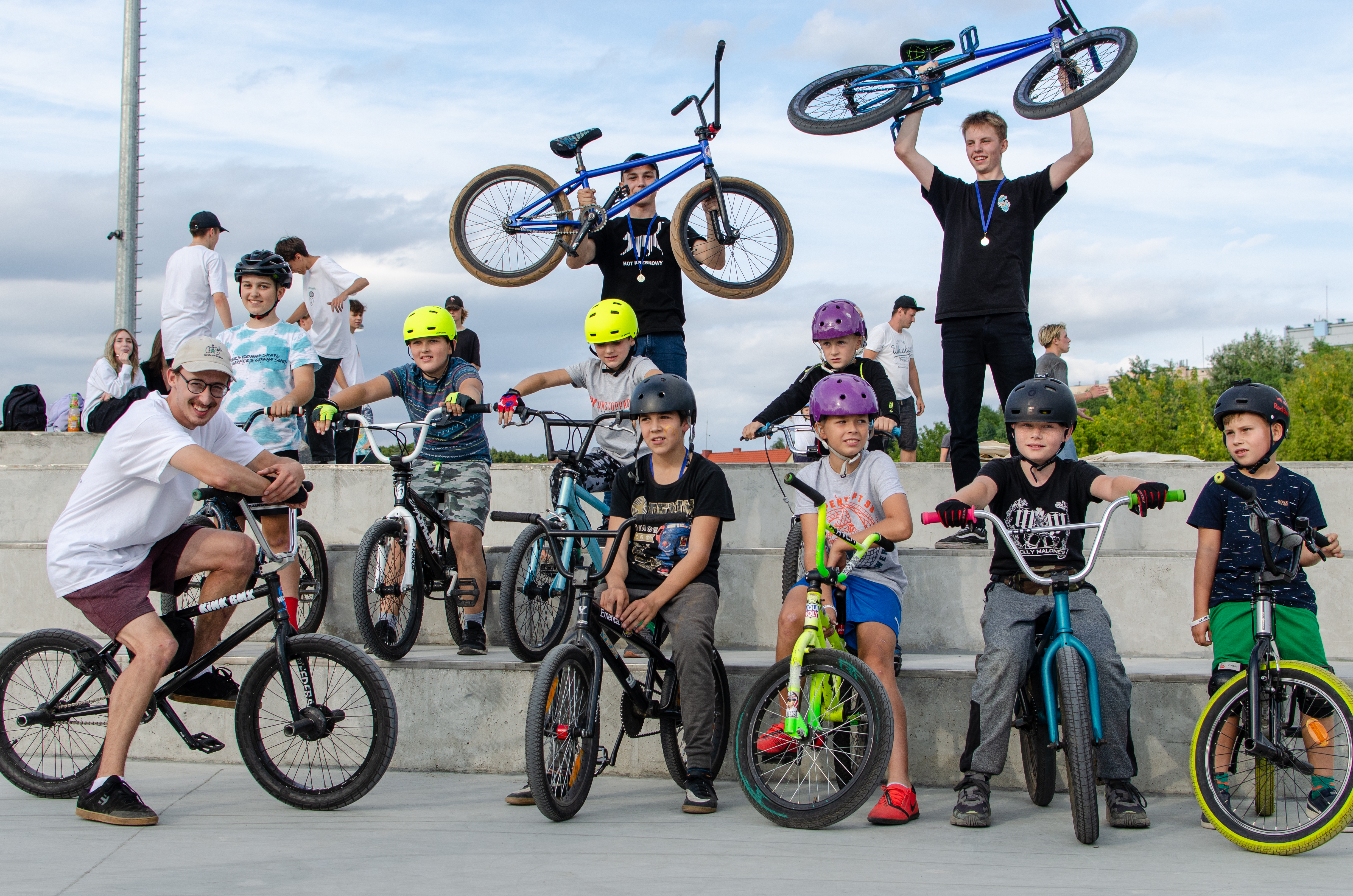 Dziewięciu chłopców siedzi na rowerach bmx. Ośmiu ma kaski na głowach, jeden czapkę basebolówkę. Za nimi dwóch chłopców swoje bmx trzyma w rękach nad głową. W tle widać kilkoro młodych dziewcząt i chłopców oraz zieleń.
