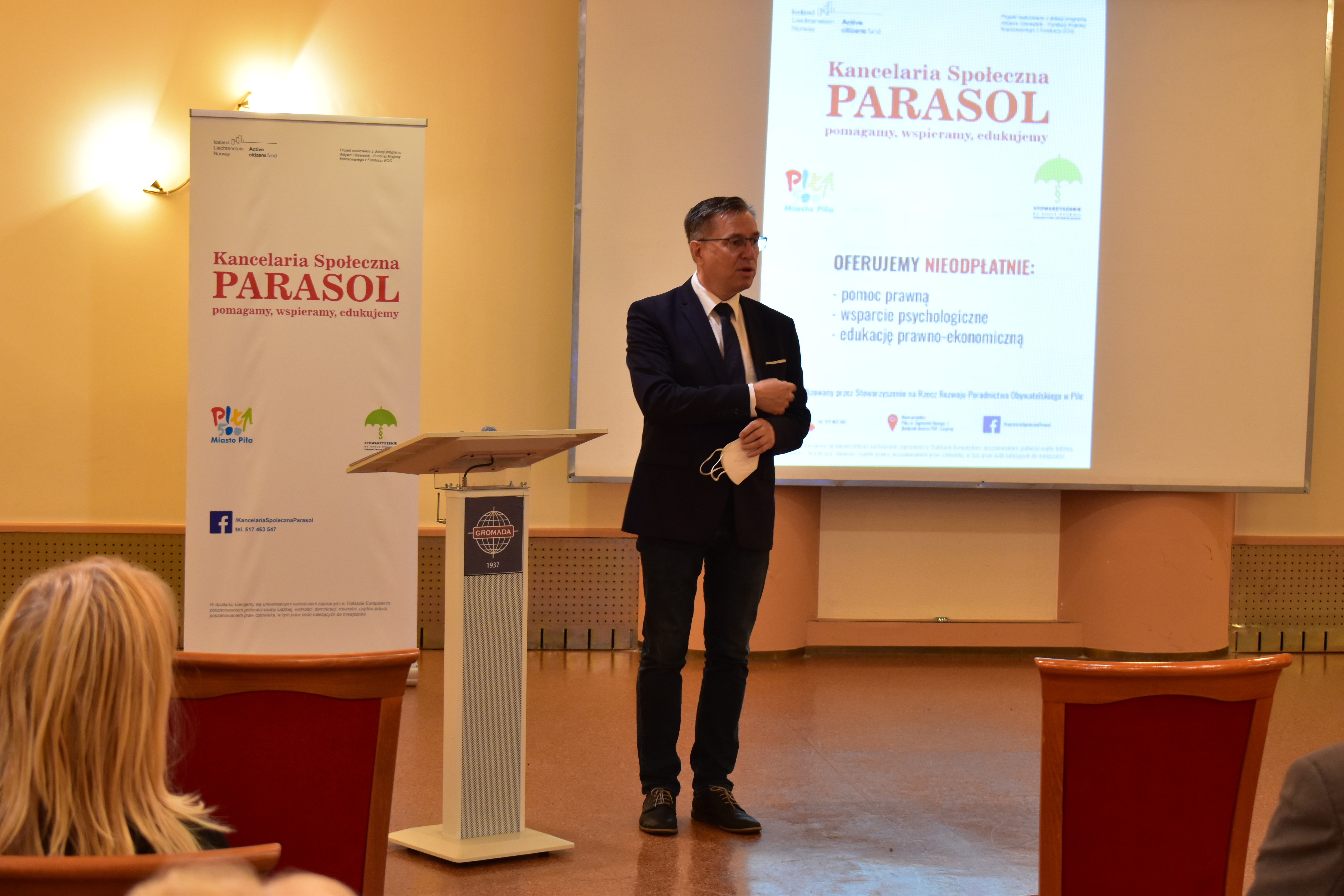 Mężczyzna w&nbsp;garniturze stoi na&nbsp;tle ekranu, na&nbsp;którym widać wyświetlony napis: Kancelaria Społeczna Parasol. Poniżej dwa logotypy: Miasto Piła i&nbsp; Kancelaria Społeczna Parasol. Poniżej informacja: Oferujemy nieodpłatnie: porady prawne, wsparcie psychologiczne, edukację prawno-ekonomiczną. Po lewej stronie mównica i&nbsp;rolap z&nbsp;tymi samymi informacjami co na&nbsp;ekranie. 