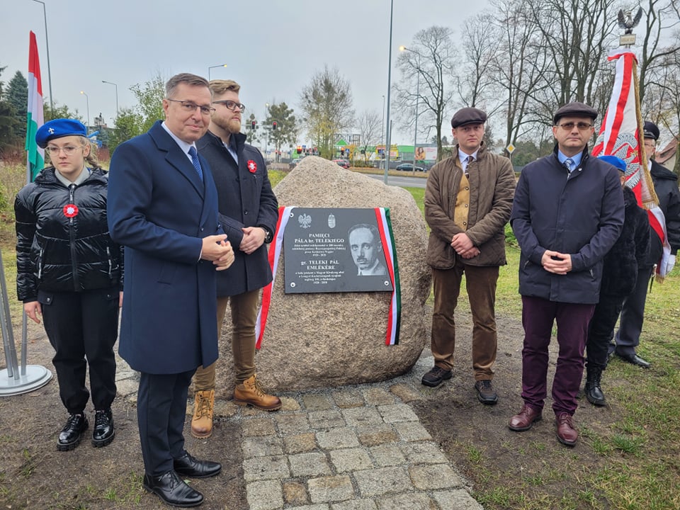Grupa ludzi stojąca na świeżym powietrzu, zgromadzona wokół pomnika - kamienia z tablicą pamiątkową, przy której wiszą flagi Polski i Węgier. Na tablicy można przeczytać napis "pamięci Pala Telekiego". Wszyscy zwróceni są w stronę fotografa.