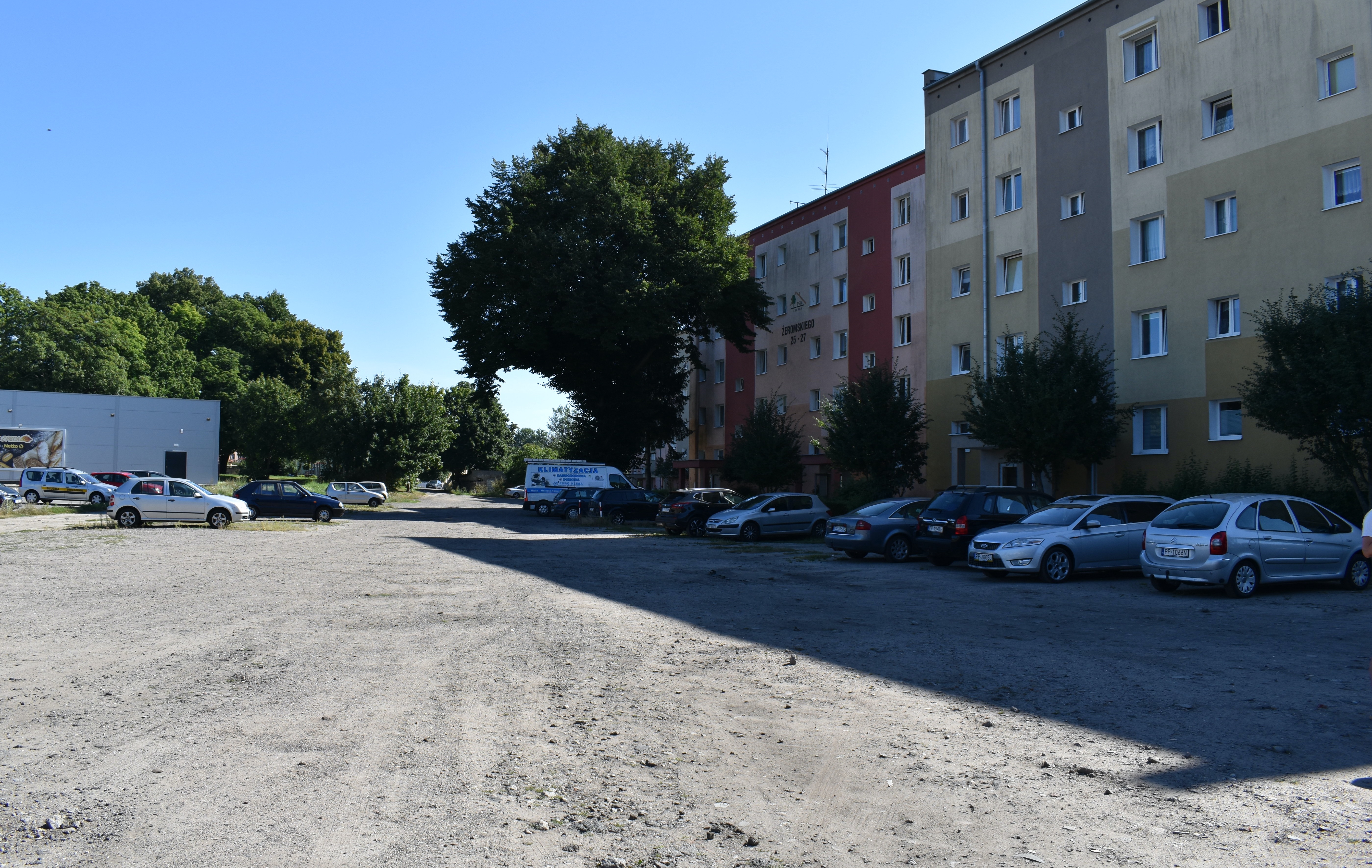 Plac piaskowo-żwirowy, po prawej stronie wielorodzinny budynek mieszkalny i&nbsp;rząd zamaskowanych przed nim samochodów. W oddali po lewej stronie budynek sklepu, tablica reklamowa przed nim i&nbsp;kilka zaparkowanych aut. W tle zieleń drzew.