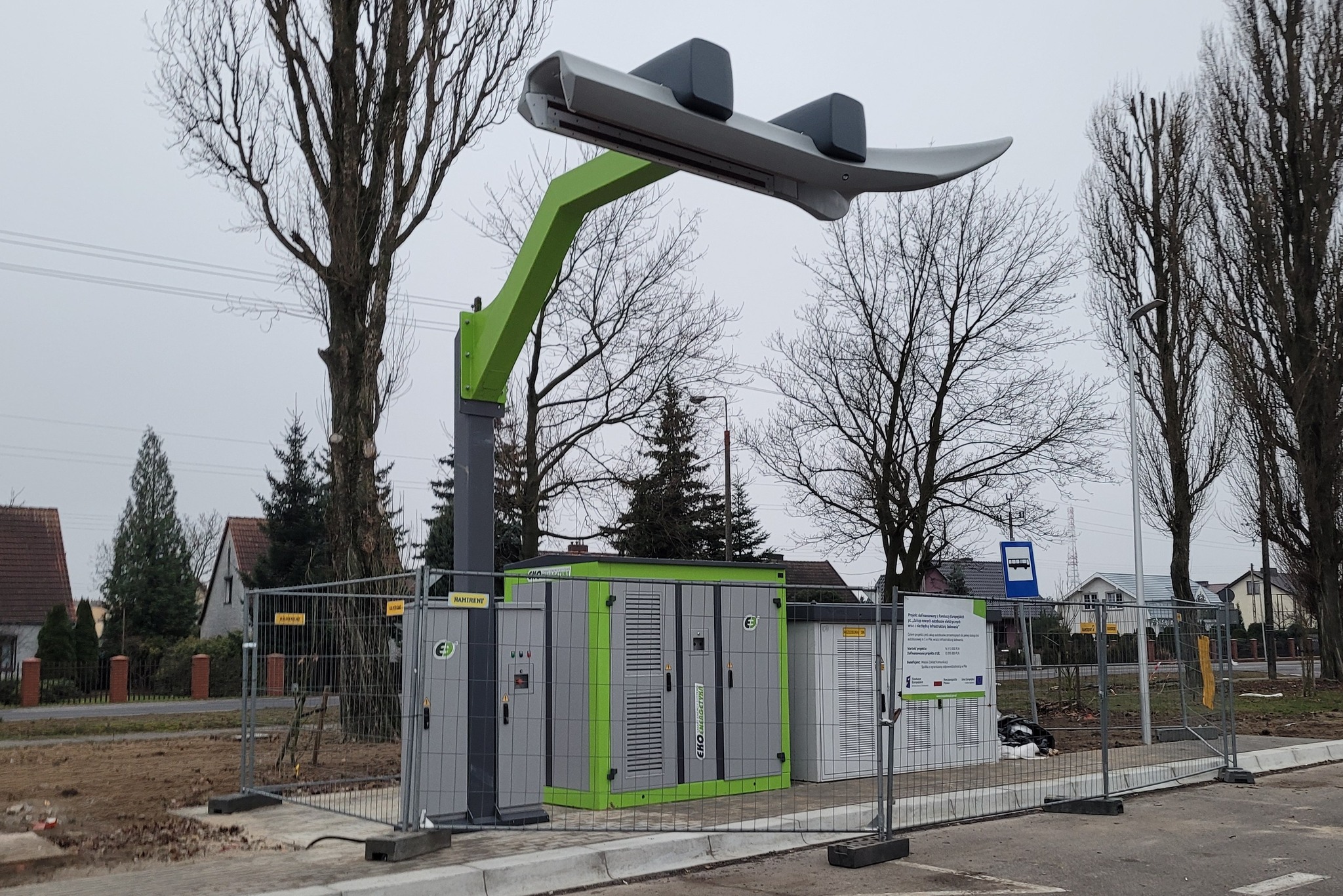 Stacja szybkiego ładowania autobusów elektrycznych podstawiona w przestrzenią miasta. Stacja jest ogrodzona metalowym płotem. W tle widać domki jednorodzinne i drzewa.