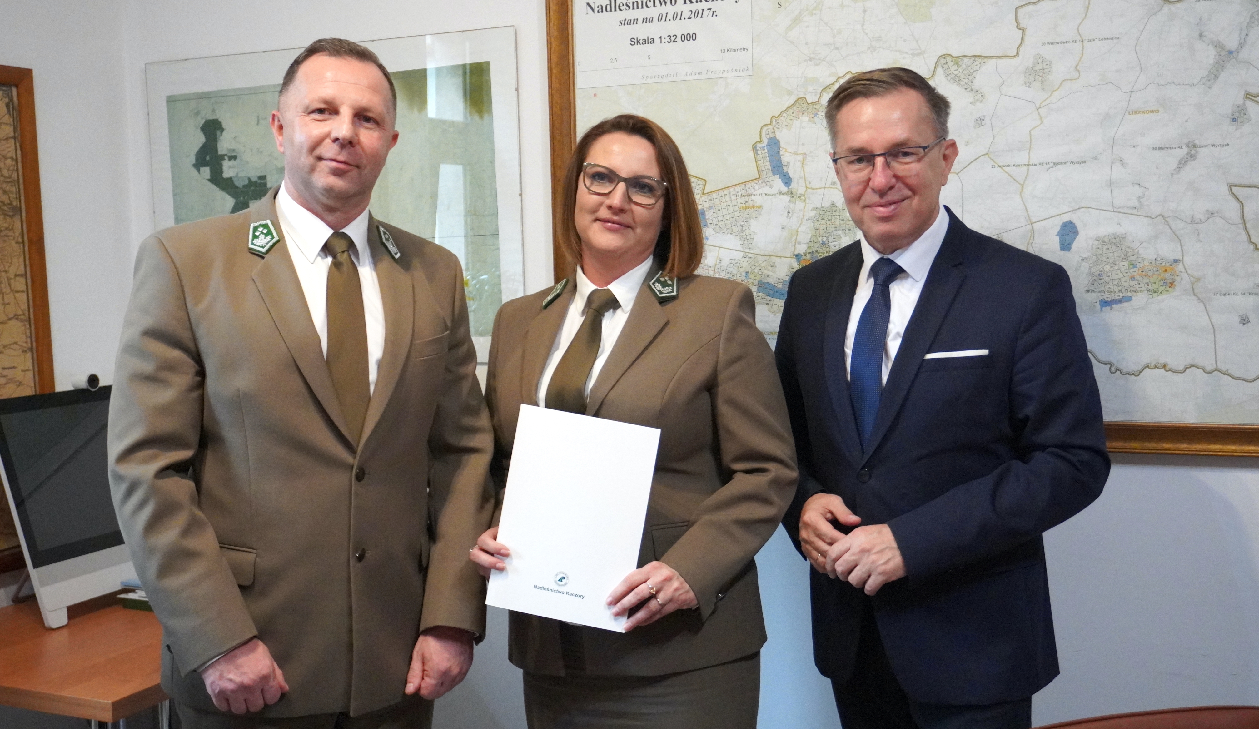Dwóch mężczyzn i kobieta w biurze, pani trzyma teczkę. Wszyscy są ubrani elegancko, kobieta i mężczyzna w mundurach nadleśnictwa. Na ścianie w tle wisi mapa.