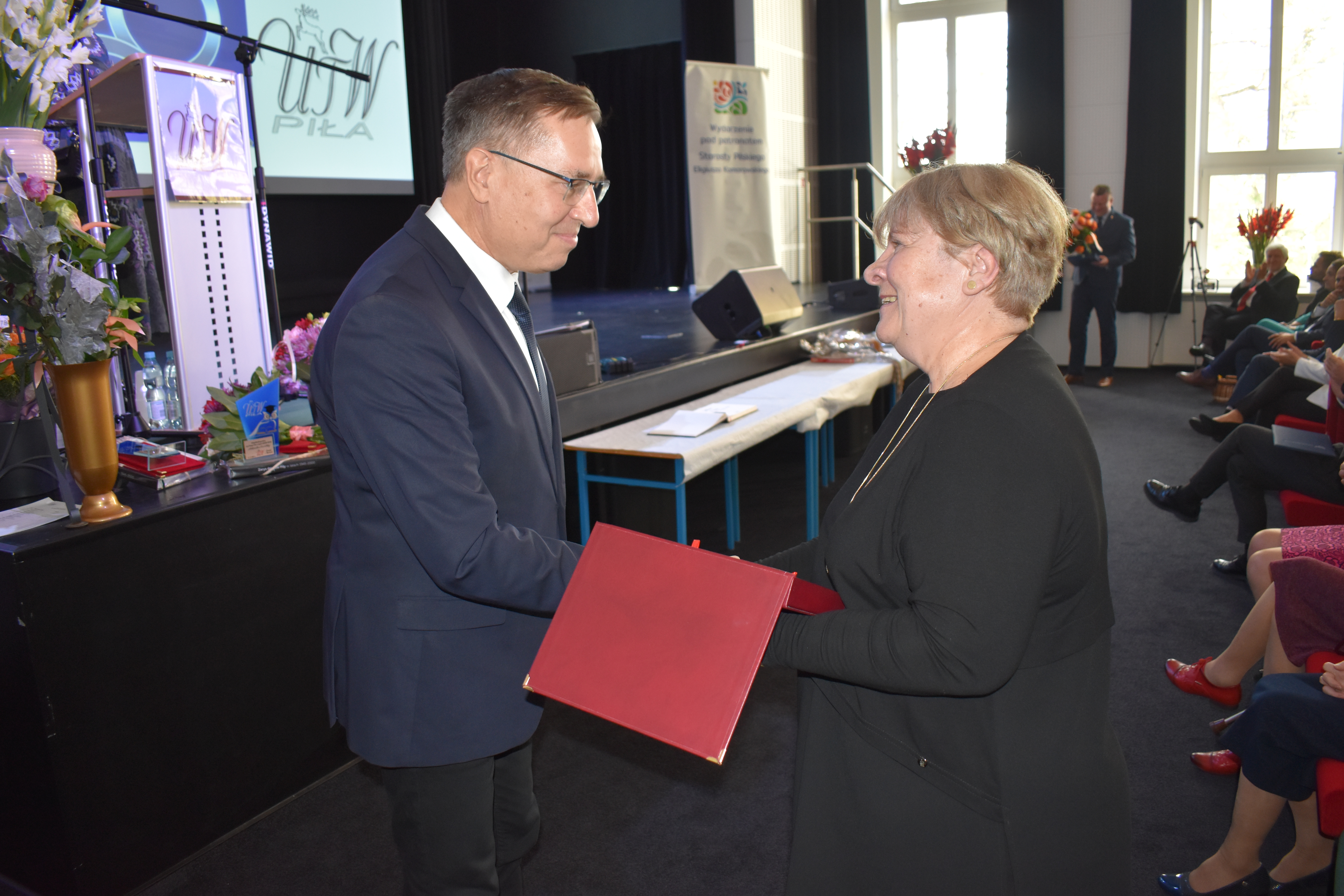 Mężczyna wręcza kobiecie pamiątkową tabliczkę. W tle ludzie siedzący w fotelach. Kwiaty.