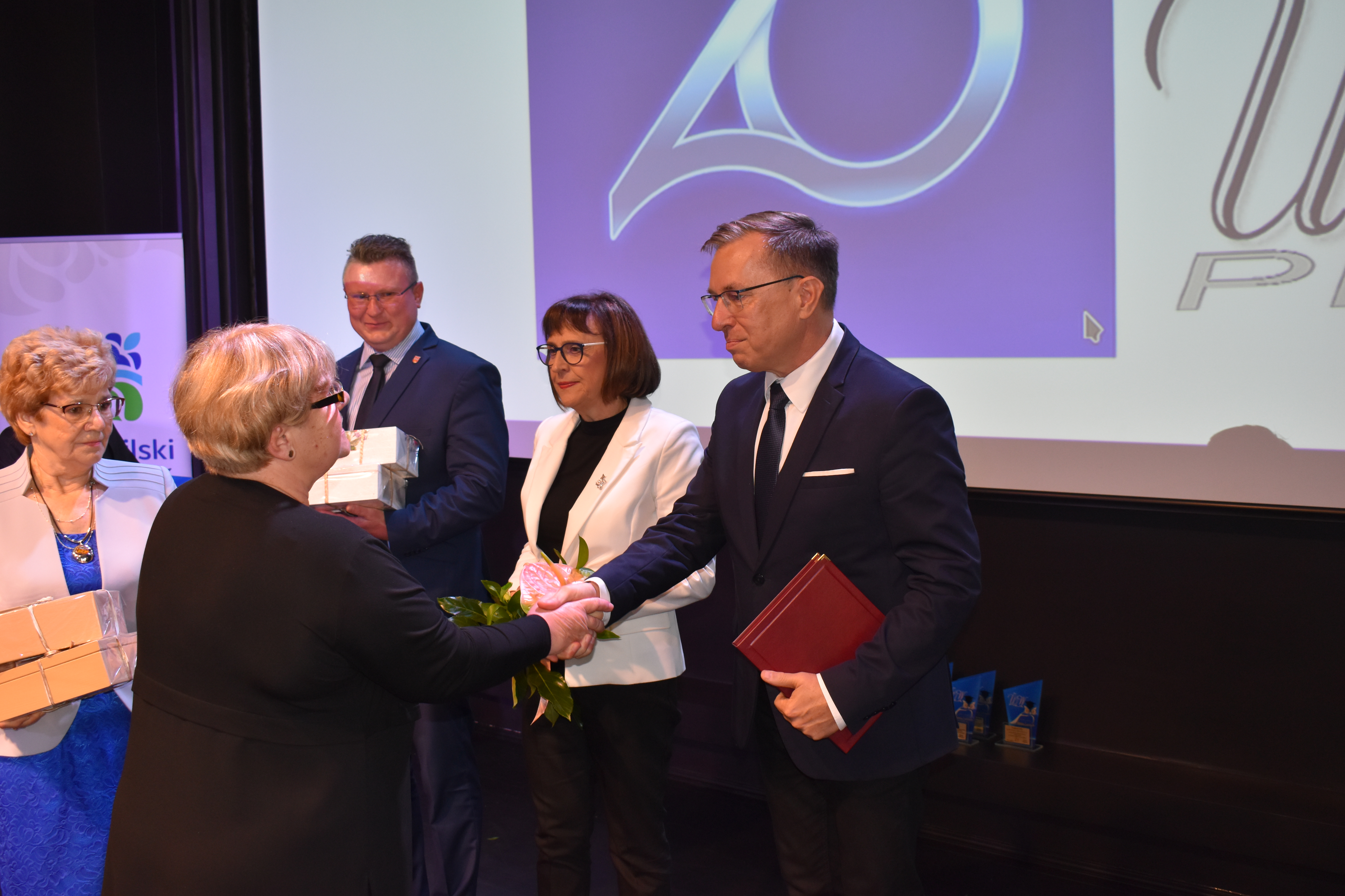 Trzy obiety i dwóch mężczyzn na scenie. Dwie kobiety wręczją kwiaty i upominki. Gratulacje. Napis 20 lat UTW w Pile.