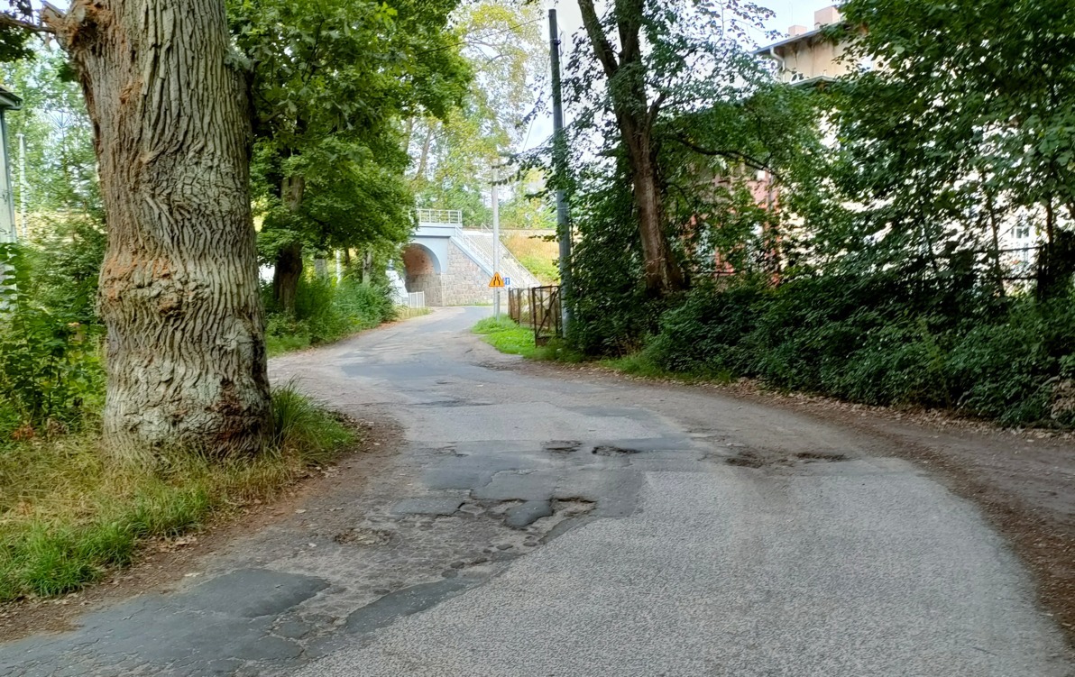 Fragment zniszczonej drogi, po dwóch stronach zieleń drzew i krzewów, zabudowania i most nad drogą.