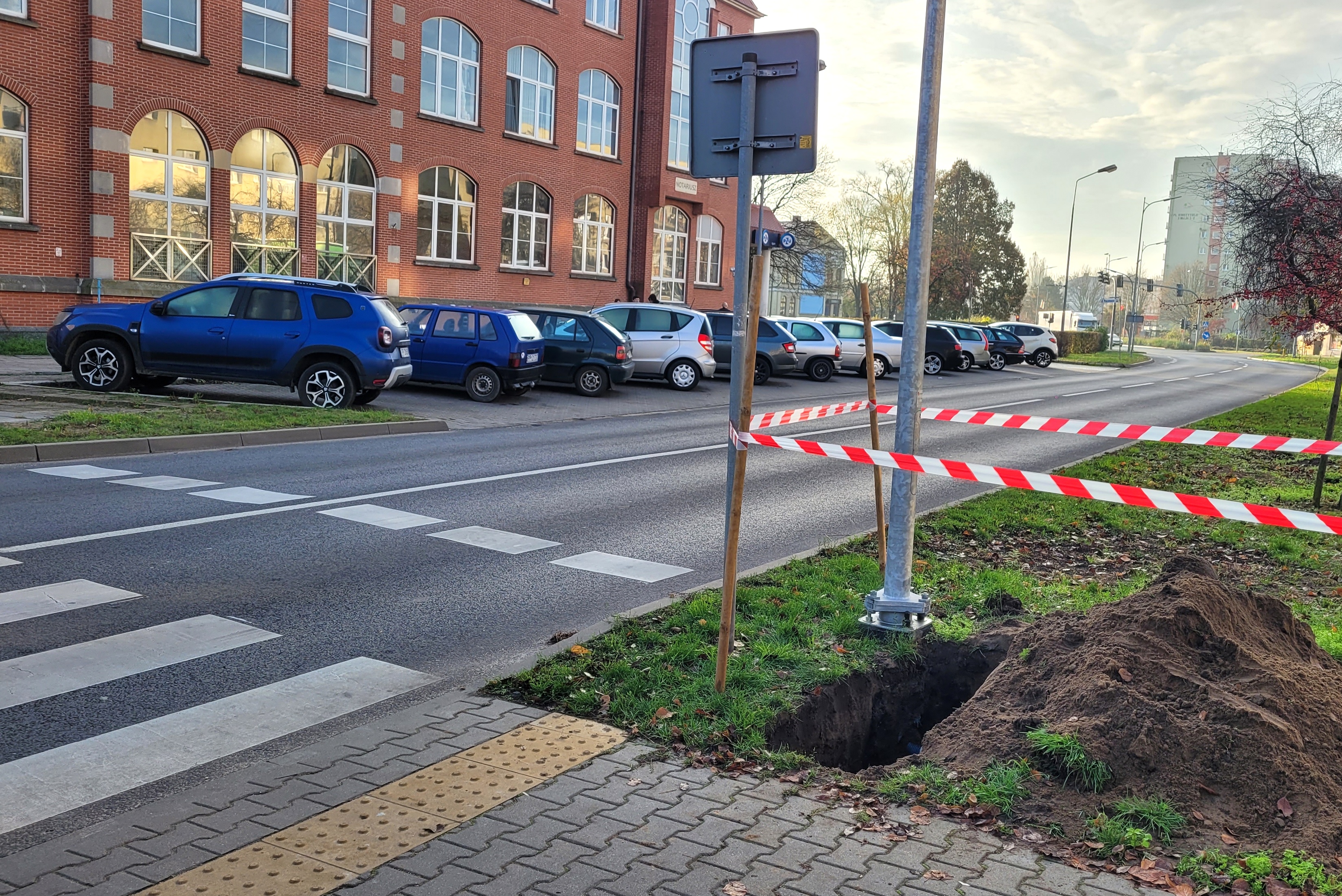 Przejście dla pieszych. Wykopany rów. Samochody ospowe na pargingu. Budynek.
