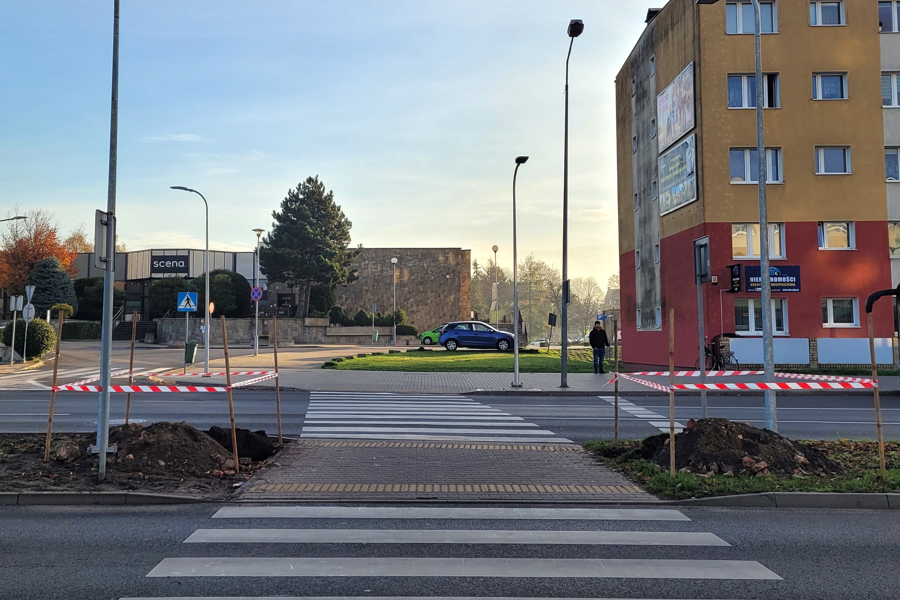 Przejście dla pieszych w mieście. Po obu stronach wyo\kopy ogrodzone biało czerwoną taśmą. W tle budynki.