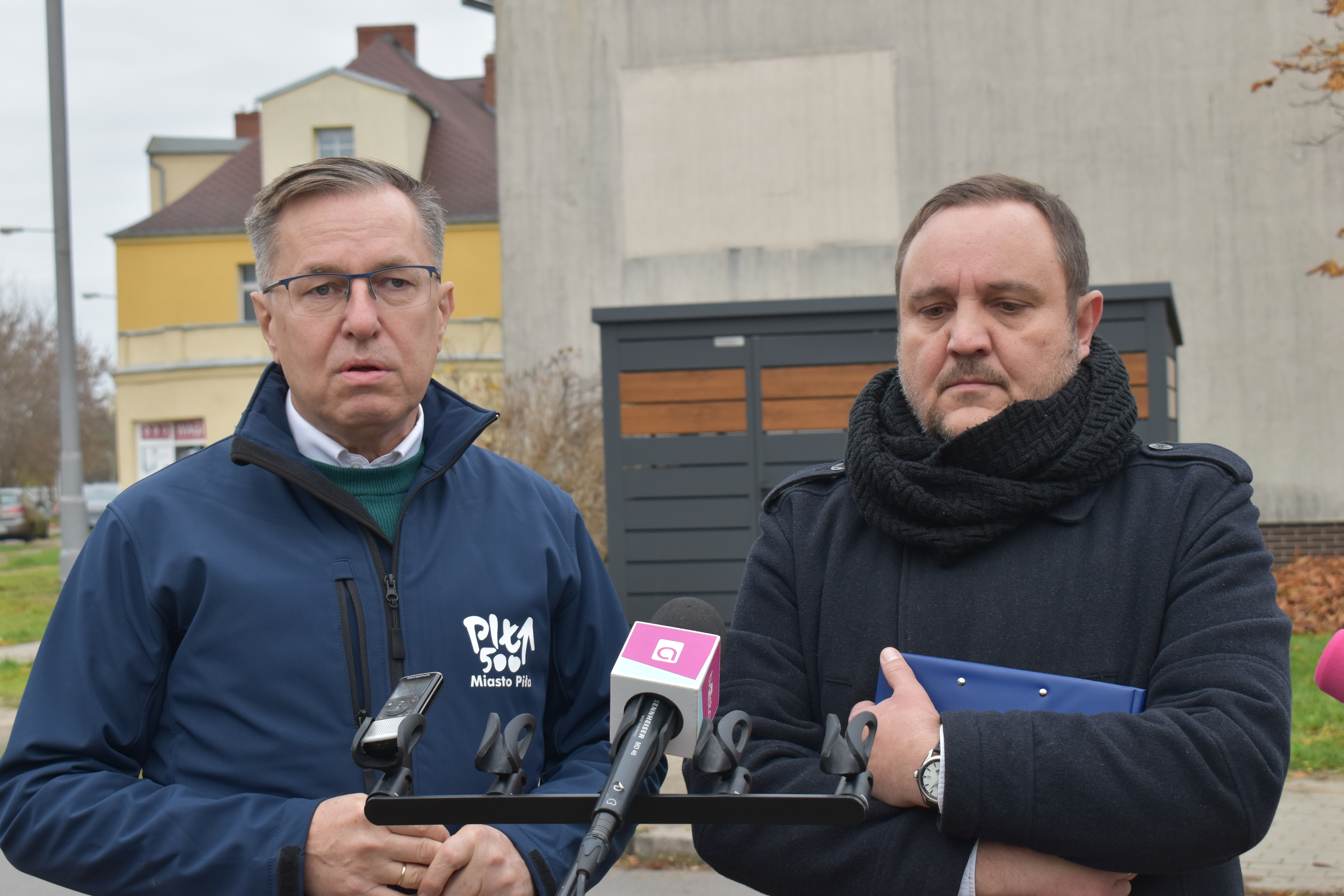 Dwaj mężczyźni stoją na ulicy przed mikrofonami. W tle budynki mieszkalne i wita śmietnikowa.