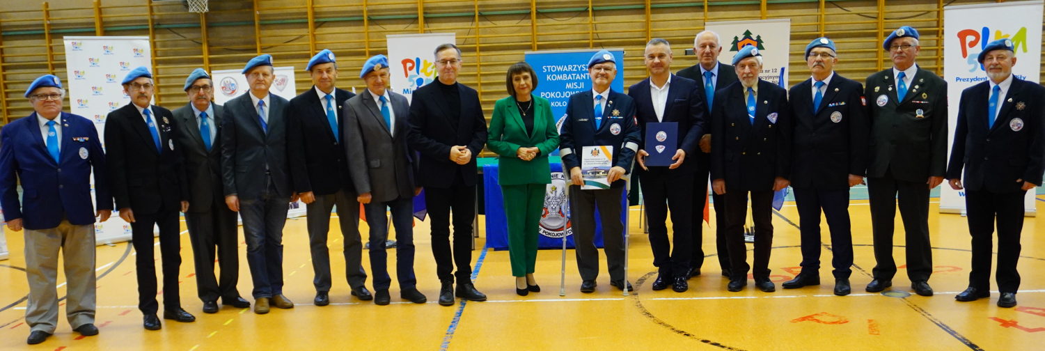 Uroczystośc w szkolnej sali gimnastycznej. Grupa kobiet i mężczyn stoi w rzędzie.