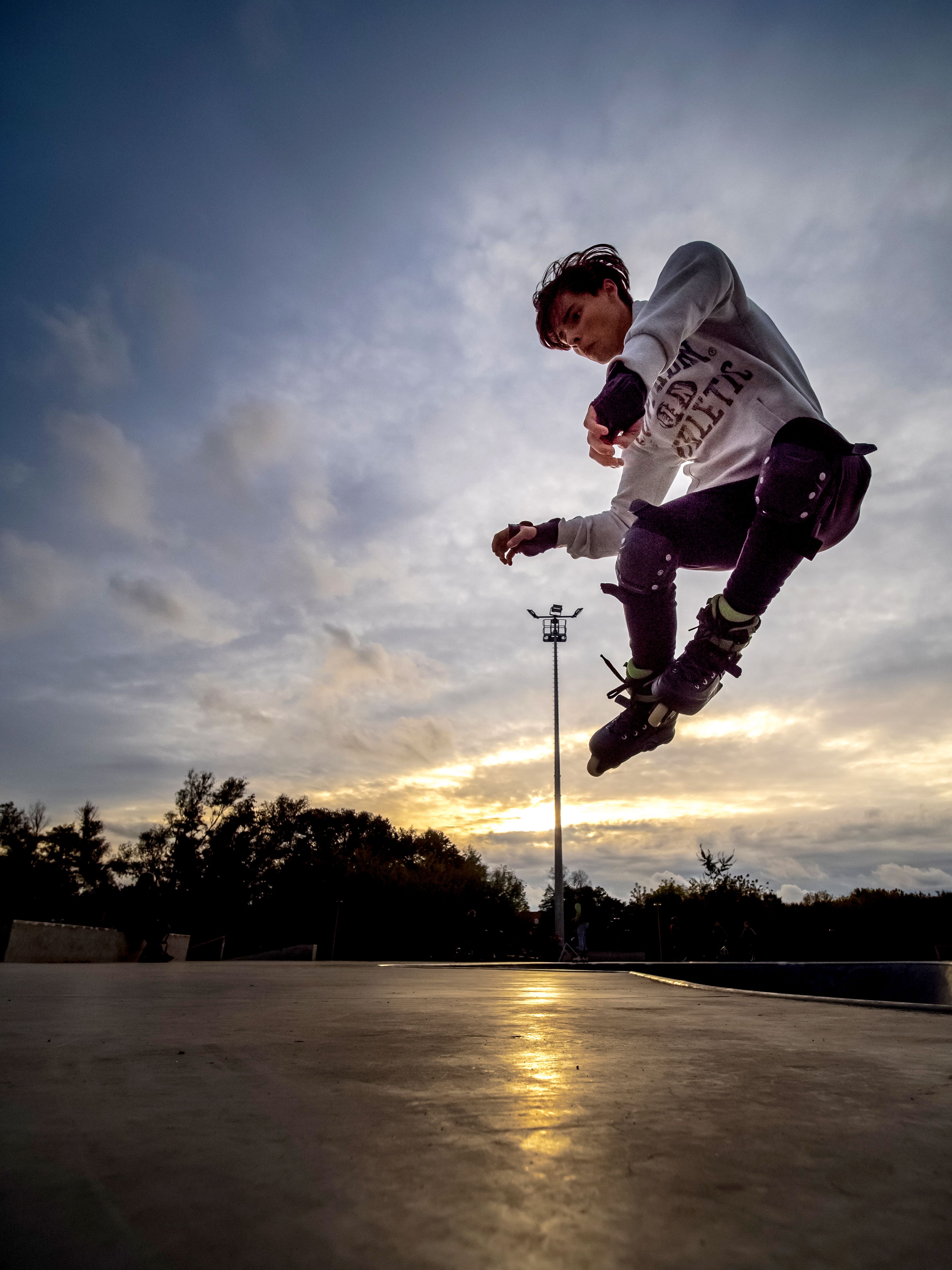 Chłopiec wykonuje ewolucje na&nbsp;rolkach hulajnodze. Ochraniacze, elementy skateplazy. Maszt oświetleniowy.   