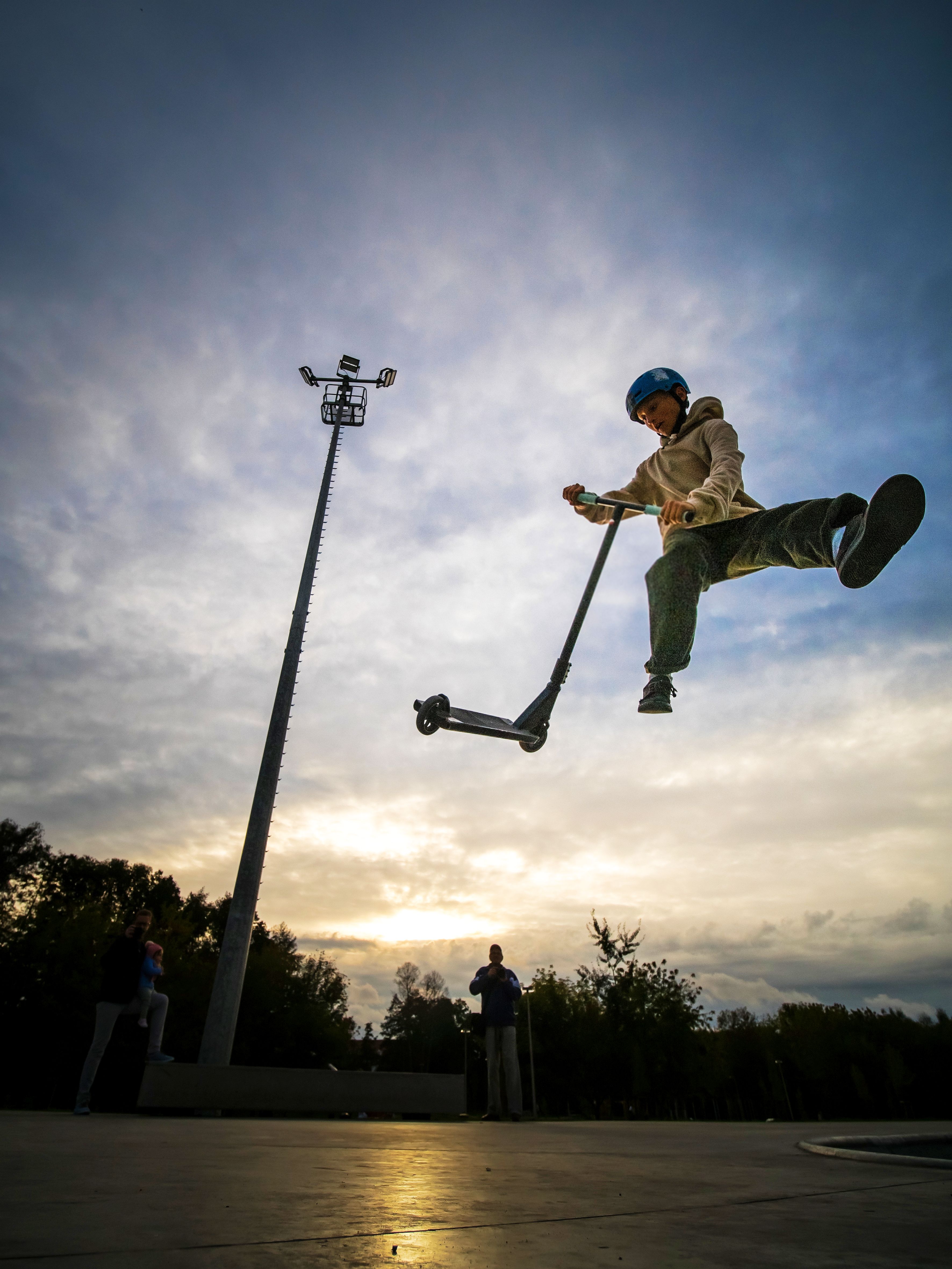 Chłopiec wykonuje ewolucje na&nbsp;hulajnodze. Kask, ochraniacze. Elementy skateplazy. Maszt oświetleniowy. Mężczyzna robi zdjęcie.  
