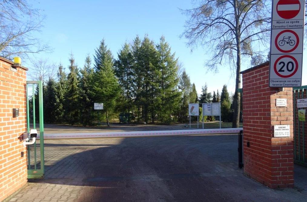 Brama wjazdowa na cmentarz, w bramie zamknięty szlaban. Po dwóch stanach mur z czerwonej cegły. Za płotem tablice informacyjne, drzewa. Po prawej stronie znak informujący o tym, że zarządca cmentarza musi wyrazić zgodę na wjazd.